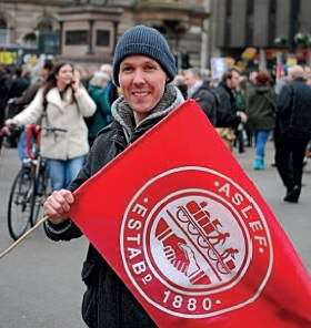 Craig Cameron is wearing a hoody, coat and warm hat. He is in a crowded street and holding a red ASLEF flag.