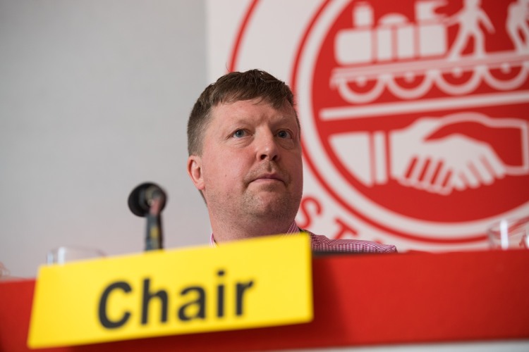 Darran Brown sits at a red desk in front of an ASLEF logo. A yellow card in front of his microphone says Chair.