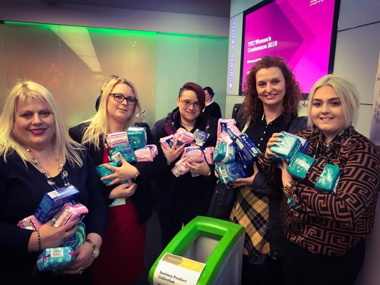 A group of five women stand around a donation box, each holding several packets of tampons and sanitary towels.