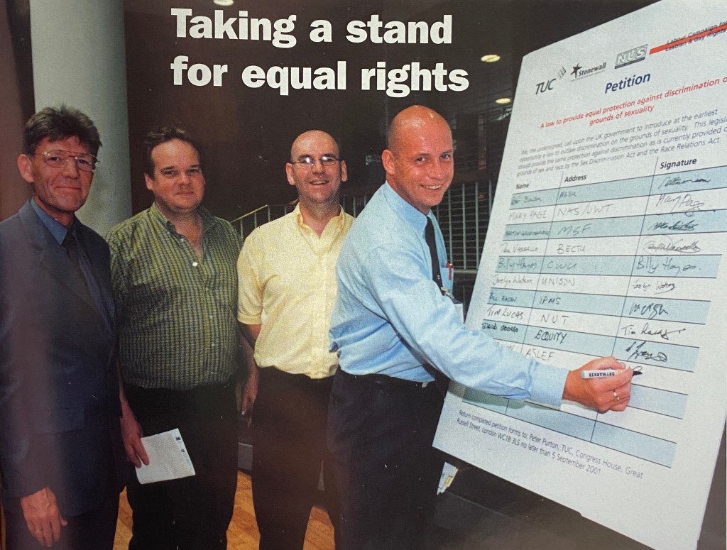 Four men stand together in a hall. On the right of the image, one of the men is signing his name on a large board that says 'petition' at the top.