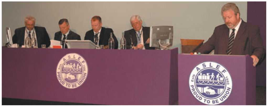 Four men sit at a purple desk which has an ASLEF logo on it. They have screens, water, and papers in front of them. On the right, a man stands at a lectern which also has an ASLEF logo on it.