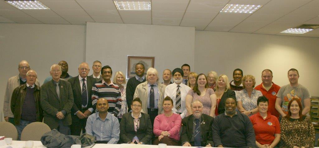 A group of about 30 people stand and sit grouped together in a room with bright lights and white walls.