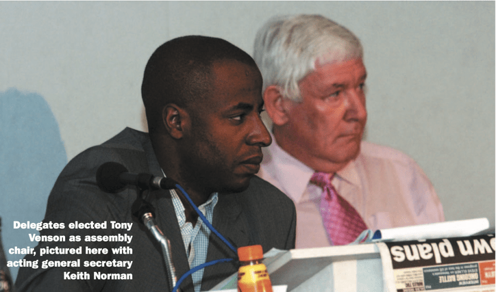 Tony Venson sits next to general secretary Keith Norman. Both have serious expressions. They have microphones and a raised desk in front of them.