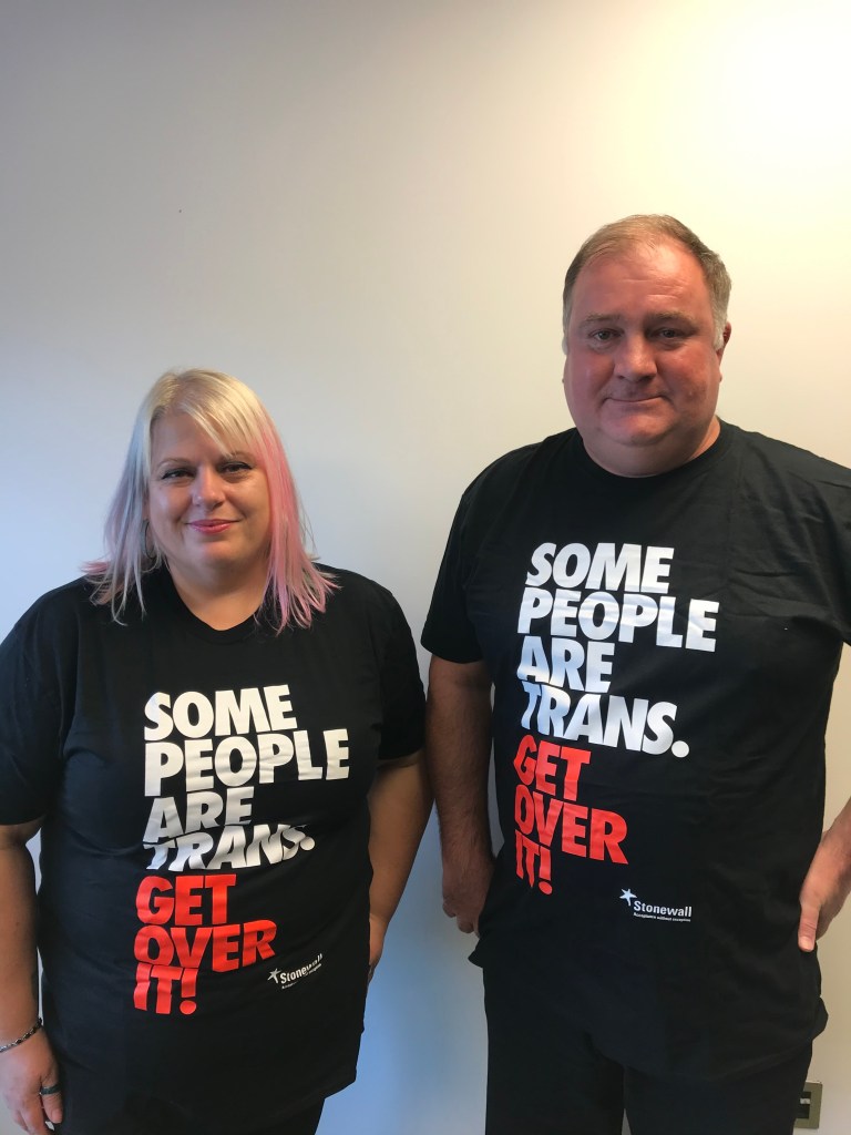 Debbie and David stand next to each other, both wearing 'some people are trans, get over it' t shirts with red writing on a black background and a Stonewall logo