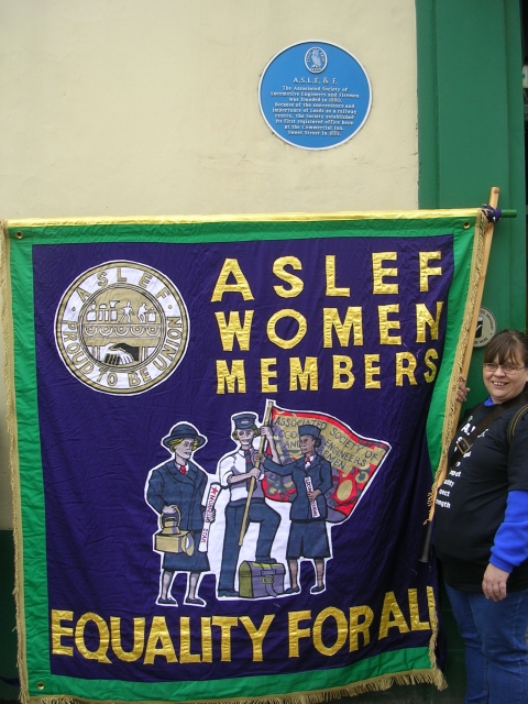A woman standing under a blue plaque which reads ASLE&F. She is holding one side of a banner which is green and purple and says 'ASLEF Women Members - Equality for All' in gold alongside a picture of three women in work uniforms